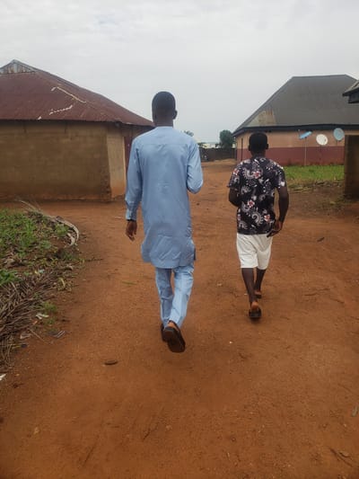 Two young Black males are walking away from the camera down a dirt path in what appears to be a rural area or the outskirts of a town. The male on the left is wearing a light blue traditional outfit, while the male on the right is wearing a black shirt with a floral pattern and white shorts. The environment consists of two buildings with corrugated metal roofs and what look like satellite dishes mounted on the side of one of the buildings. The ground is reddish-brown dirt, and there is some vegetation along the left side of the path. The sky is overcast.