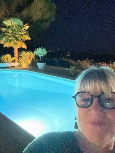 The image shows a woman with blonde hair and glasses taking a selfie at night by a swimming pool. The pool is illuminated with blue light, and a palm tree and other plants are visible in the background, also lit by artificial lights. In the distance, city lights can be seen. The sky is dark with some stars. The woman is wearing a dark top and large, dark-rimmed glasses. She has earrings on. The scene suggests an outdoor setting, possibly a resort or a private residence with a swimming pool and garden. There is no specific indication of the city of Saint-Galmier, France, in the image itself.