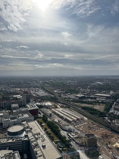 This is an aerial photograph looking northeast over a section of Birmingham, United Kingdom. The foreground features a cluster of modern buildings, some with large, flat roofs adorned with numerous HVAC units and other rooftop infrastructure. One building in the foreground has a distinct circular glass structure on its roof. Adjacent to these modern structures, there is a large construction site with visible scaffolding, excavation, and building materials.
Further into the mid-ground, a railway yard with multiple tracks and platforms is visible, appearing to be undergoing some form of development or renovation. Several long, covered structures stand over the platforms. Beyond the railway yard, the city stretches out into the distance, characterized by a mix of residential and commercial buildings interspersed with trees. A major railway line runs through the cityscape, with trains visible on the tracks. The horizon is hazy, suggesting a considerable distance to the furthest visible urban sprawl.
The sky above is partly cloudy, with the sun shining through breaks in the clouds, creating bright patches and casting a diffused light over the scene. The overall impression is one of a developing urban landscape under a bright, yet somewhat overcast sky. No people are discernible in the image.
