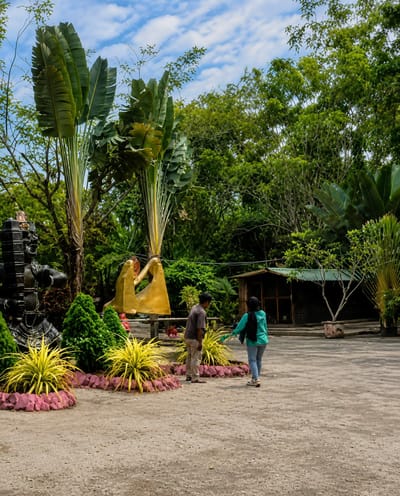 The image depicts an outdoor scene at what appears to be a tourist attraction or park in Kota Pari, Indonesia. In the foreground, there is a paved area with loose gravel. To the left, a large, dark, ornamental sculpture is visible. In the center of the image, two people are interacting. A person wearing a brown shirt and khaki pants is standing with their back partially turned to the camera, looking towards a second person. The second person, wearing a teal hijab and light blue jeans, is facing away from the camera and appears to be gesturing or pointing towards something on the left. Between the people and the sculpture, there are several plants, including brightly colored yellow foliage plants with pinkish-red bases, and several cone-shaped green bushes. Behind these, two tall, fan-like palm trees dominate the left side of the background. Further back, a small wooden building with a green roof is situated. Lush green foliage and trees form the backdrop of the scene, with a cloudy blue sky overhead.