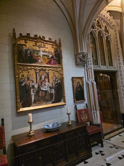 The photograph captures an interior view of a Gothic-style room, likely within a historical building. Dominating the scene are two large, ornate religious paintings displayed on a pale, textured wall. The upper painting depicts the crucifixion of Jesus, with figures of Mary and John present. The lower painting shows the Virgin Mary enthroned with the infant Jesus, surrounded by saints. These paintings are framed in elaborate gold leaf.
In the foreground, a dark, intricately carved wooden chest of drawers serves as a display surface. On top of the chest are two brass candlesticks with lit candles and a blue and white ceramic bowl. To the left of the chest, part of a red upholstered chair is visible. To the right of the chest, a separate, smaller framed painting of the Madonna and Child hangs on the wall.
The architecture features pointed arches, ribbed vaulting, and decorative stonework. To the right, a grand arched doorway leads into another room, partially obscured by a glass barrier. Stained glass windows, depicting figures in armor, are visible through the arched opening of the doorway. The floor is a black and white checkered pattern.
The scene is static, with no visible individuals actively engaged in any interactions within the frame, although figures are depicted within the paintings. The lighting appears to be a mix of natural light filtering through windows and artificial light from the candles and possibly unseen sources, casting shadows and highlighting the details of the artwork and furnishings.
Based on the architectural style and the presence of religious art, this location is likely a museum or a historically significant religious building. However, without further context or external information, it is not possible to definitively place this scene within a specific location in Utrecht, Netherlands. The image solely depicts the interior of this room.
