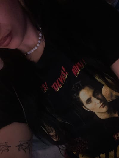 This close-up, dimly lit, indoor photograph depicts a female subject from the neck downwards to the upper thigh, located in Saint-Leu-la-Forêt, France. The subject wears a black t-shirt featuring a graphic print with the words "WOLVES! ROAR!" in red and yellow text, alongside an image of a male face with dark hair and freckles. A choker-style necklace composed of light-colored spherical beads interspersed with smaller metallic beads adorns her neck. Long, dark hair is visible, draped over her left shoulder and chest. On her lower left arm or upper left thigh, a black outline tattoo depicting two stylized bat or dragon wings is present. The background is dark and out of focus, indicating an interior setting with limited ambient light.