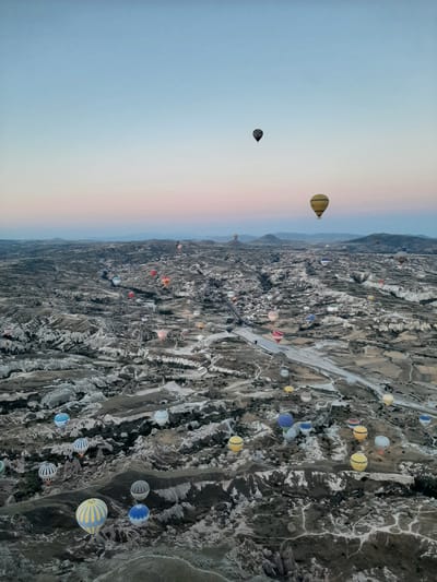 This aerial photograph captures a landscape dotted with numerous hot air balloons in Cappadocia, Turkey. The scene depicts a vast, rugged terrain characterized by unique geological formations, including valleys and rock chimneys. The sky is a gradient of soft blues and pinks, indicating either dawn or dusk. The balloons, in various colors and patterns, are scattered across the frame, some ascending and others drifting at different altitudes. The image offers a sense of scale and wonder, highlighting the popularity of hot air ballooning as an activity in this region.
