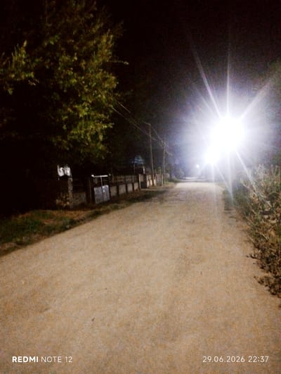 This is a nighttime image of a dirt road in the city of Ak-Suu, Kyrgyzstan. The road is unpaved and appears to be a rural or semi-rural area. On the left side of the road, there is dense foliage from trees and bushes. Further down the road on the left, there are structures that appear to be fences and possibly buildings. On the right side of the road, there is also vegetation and a ditch.
The dominant feature in the image is a bright, intense light source, possibly a streetlight, located in the upper right quadrant of the image. This light creates strong lens flare and illuminates the road and the surrounding environment. The light is so bright that it washes out details in its immediate vicinity and creates stark shadows.
The image includes superimposed text in the lower left corner which reads "REDMI NOTE 12" and in the lower right corner which reads "29.06.2026 22:37". These are likely camera model and timestamp information.
There are no people or animals visible in the image. The scene is still and quiet, with the primary visual element being the contrast between the darkness of the night and the intense artificial light. The overall impression is one of a deserted road illuminated by a powerful light source.
