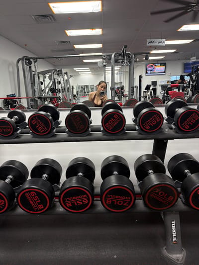 The image shows a gym interior in Las Vegas, United States. A person stands in front of a wall of mirrors, holding a phone, presumably taking a selfie. The gym contains various weightlifting machines and equipment. Foreground focus is on a dumbbell rack with weights ranging from 10 to 70 lbs. A TV screen is visible on the wall, as well as a sign reading "Pain 2 Gain".