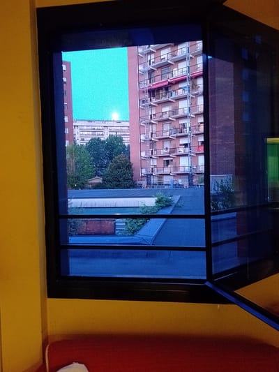 The image, taken from an interior vantage point looking out a window, depicts an urban scene in Turin, Italy, characterized by multi-story apartment buildings and a clear sky. The window frame, dark and imposing, divides the view into distinct sections, with the right side of the frame revealing a slightly open window and a hint of a green tint, possibly from a reflection or tinted glass.
In the background, a tall, salmon-pink apartment building dominates the right side of the frame, its numerous balconies adorned with railings and some featuring red awnings. Another similar building is partially visible on the left, behind a band of trees. The sky above is a vibrant, almost electric blue, with a subtle glow in the distance suggesting either the moon or a strong light source. Below the buildings, a flat, dark roof surface is visible, interspersed with patches of greenery. Further down, a red brick structure with a corrugated metal roof is discernible, hinting at storage or utility areas. The foreground, viewed through the lower pane of the window, is a deep blue, reflecting the sky or the window's tint.
The overall impression is one of a residential urban environment, likely taken during daylight hours, possibly late afternoon or early evening, given the quality of light. The composition, framed by the window, creates a sense of looking into another space, a common perspective from a residential apartment in a city like Turin. There are no individuals or any discernible human activity captured within the frame. The scene is static and purely observational.