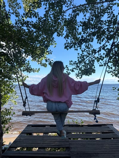 A blonde woman wearing a pink hoodie and jeans sits on a wooden swing suspended from tree branches overlooking a body of water. She is positioned on a weathered wooden platform near the water's edge. The sky is blue with scattered clouds, and the water is choppy. The environment suggests a natural, outdoor setting in the vicinity of Podstepki, Russia.