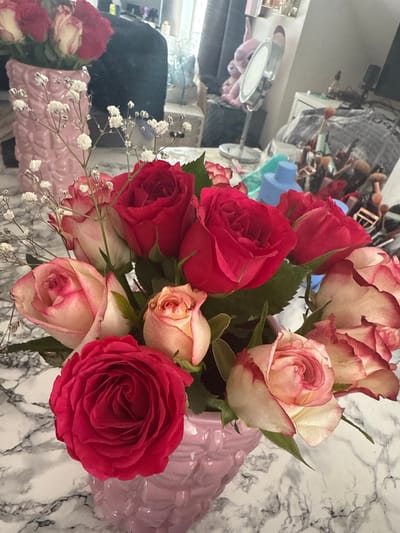 This image depicts a floral arrangement situated on a white and gray marble-patterned surface, presumably a countertop or table, in a residence located in Vellmar, Germany. The primary subject is a bouquet of approximately 15-20 roses, presented in a light pink, textured ceramic vase. The roses are a mix of vibrant red and bi-color pink-and-white varieties, interspersed with delicate white baby's breath.
In the blurred background, reflected in what appears to be a mirror or glossy surface, is a cluttered interior space. Visible objects include a second, similar pink vase containing more roses, various cosmetic items such as makeup brushes in containers, bottles, a standing vanity mirror, and a pink plush toy resembling "Angel" from Disney's Lilo & Stitch. The overall environment suggests an indoor setting, likely a bedroom or dressing area, with diffuse lighting. No individuals are present or directly discernible in the image.