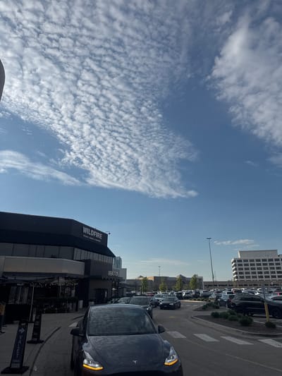 The image, captured in a portrait orientation, depicts a vibrant outdoor scene in a commercial area, likely Oak Brook, USA, under a bright daytime sky. The upper and left sections are filled with a striking blue sky adorned with dense patterns of white, altocumulus-like clouds. The clouds vary from tightly clustered formations to more wispy, scattered elements.

On the right side, a busy road or parking lot is visible, featuring numerous parked cars, predominantly dark in color, and clear white striped road markings, resembling a crosswalk. In the immediate lower right foreground, a dark vehicle, possibly a Tesla, is prominent, its front right turn signal illuminated.

The lower half of the image showcases several modern buildings. A large, multi-story structure with numerous windows, potentially an office building or parking garage, rises in the background. Closer to the viewer, a dark-colored building prominently displays "WILDFIRE STEAKS, CHOPS & SEAFOOD" in white lettering on its side. Nearby, another establishment is identified as "BEATRIX." Outdoor dining areas with string lights are visible below these establishments. Valet parking is indicated by signs, including one that reads "VALET PARKING" and another, "VALET CLOSED Thank you," positioned near the foreground vehicle, suggesting an upscale dining and retail environment. The clear blue sky and bright, even lighting confirm it's a sunny day with fair weather. No specific human activity or emotions are discernible.
