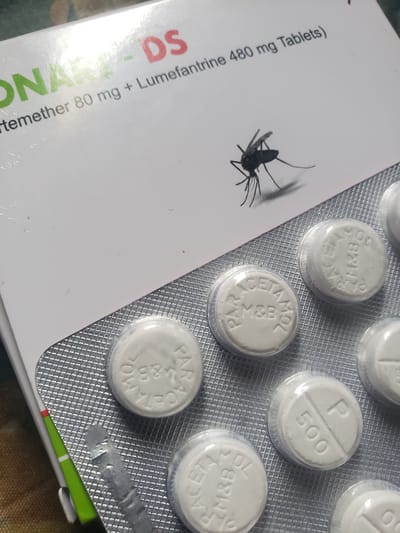 The image shows a blister pack of white round tablets, some of which are partially visible. The packaging features text that includes "ARTEMETHER 80 mg + Lumefantrine 480 mg Tablets". There is also a graphic of a mosquito on the packaging. The tablets themselves have markings, with some showing "PARACETAMOL M&B" and others showing "P 500". The blister pack is positioned on a surface that appears to be a table. The image does not contain any information about the city of Iganmu, Nigeria or any details related to people, actions, or interactions.