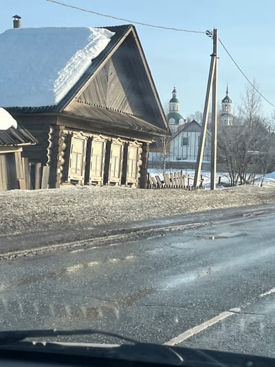 The image depicts a winter street scene in Чутырь, Russia, viewed from inside a vehicle. In the immediate foreground, the lower portion of a vehicle's dashboard and windshield wipers are visible. The asphalt road surface is wet, dirty, and shows tire tracks, with patches of ice or melting snow and a faint white dividing line.
In the midground, a traditional, weathered wooden log house (izba) occupies the left side. Its steeply pitched roof is heavily covered with snow, and a small chimney protrudes from it. The house features a facade with five tall, narrow rectangular windows, each with ornate wooden frames. A low, irregular wooden fence partially encloses the property in front of the house. To the right of the house, a wooden utility pole with overhead wires stands vertically. The ground around the house and fence is covered with dirty snow and bare earth.
In the background, under a clear blue sky, two distinct Orthodox church structures are visible. The left church features a green dome atop a white drum, capped with a golden cross. The right church presents a darker dome and a multi-tiered bell tower, also topped with a cross. Between the utility pole and the churches, bare deciduous trees and glimpses of other buildings, including a white-sided house, are present. No people or animals are visible.