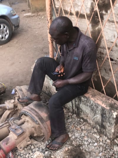 A man in dark clothing and sandals kneels among automotive parts and rubble, looking at a cell phone in his hand. He rests one foot on one of the parts. A car's wheel and the edge of the vehicle are visible on the left. A rusty metal grid is in the upper left corner of the frame. Pebbles and the edge of a concrete structure fill the upper right corner.