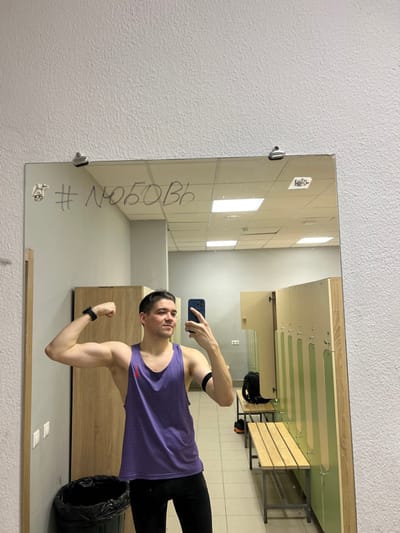 A man is taking a mirror selfie in what appears to be a locker room. He's wearing a purple tank top and black athletic pants, flexing his arm for the camera. The mirror is mounted on a wall with the hashtag "#любовь" written on it. The reflection shows lockers, benches, and other changing room elements. He is holding an iPhone in his left hand.