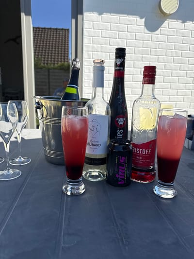 This media file captures an outdoor scene on a bright, sunny day, likely a patio or backyard, prepared for a casual social gathering. A grey plastic table is laden with an assortment of beverages and glasses.
From top to bottom in the image's orientation, two tall glasses hold a red, iced drink. Adjacent to them is a clear bottle of KRIStoff Red vodka liqueur, featuring prominent labels that read "RED - BERRY FLAVOURS & VODKA LIQUEUR" and "THE LAND OF THE WOLF", along with "ESTO 1306". Below this, a black bottle with "woodberries" on its label stands next to a black "VIRUS" energy drink can, which states "the Energy Drink planet" and "Energy Drink Guarana+".
Further down the table, a white wine bottle is clearly labeled "2025 Madame DUBARD TENDRESSE" and "DOUX & FRUITÉ". Partially nestled within a silver ice bucket is a green bottle, likely sparkling wine or champagne, with a small yellow detail on its neck. The polished surface of the ice bucket reflects a clear blue sky and a building with a dark-tiled roof. Two empty, elegant champagne flutes, bearing faint, partial brand text (e.g., "GNE NGER"), are also visible on the table.
The bright sunlight casts strong shadows, indicating it is daytime. A white tiled surface or wall forms a significant part of the background, enhancing the outdoor ambiance. The arrangement of drinks and glassware suggests an informal celebration or a relaxed get-together.