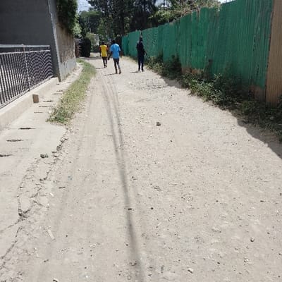 The image displays an unpaved dirt and gravel road in Ongata Rongai ward, Kenya, under bright daylight conditions. Three individuals are walking away from the camera, progressing into the background. The leftmost person wears a yellow t-shirt and dark trousers. The middle person wears a light blue t-shirt and dark lower garments, with light-colored footwear or bare feet. The rightmost person wears a dark long-sleeved shirt and dark trousers.

On the left side of the road, a concrete sidewalk or drainage channel runs parallel, bordered by a black metal fence. Behind this fence, a dark-colored building wall is partially visible, with trees and dense green foliage extending above it. On the right side of the road, a long, tall green corrugated metal fence, exhibiting signs of wear and rust, runs along the path, with small weeds and sparse vegetation at its base. The road surface is uneven, consisting of loose soil, small stones, and visible tire tracks. A distinct, elongated shadow from an unseen overhead object crosses the foreground of the path.