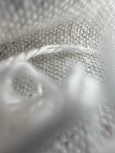 This is a close-up, abstract macro photograph of a white, knitted fabric. The texture of the knit is highly detailed, with visible loops and stitches creating a dense pattern. A strand of white yarn is prominently featured, running vertically through the frame and appearing slightly fuzzy. The lighting creates shadows and highlights on the fabric, giving it a dimensional quality. The image is almost entirely monochromatic, relying on shades of white and gray. There are areas of soft focus and blur, particularly in the upper left portion of the image, which obscures some details and adds an ethereal quality. No people, specific objects, or a discernible scene are present; the focus is purely on the texture and form of the textile. The image is abstract and does not convey a specific activity, time of day, weather, or location cues. There is no visible text.
