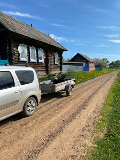 The image depicts a rural scene, likely in Kady-Sal'ya, Russia, based on the location request. A silver car with a trailer is parked on a dirt road, next to a traditional wooden house with blue-trimmed windows. A person, possibly a young man, is sitting in the trailer. The road is lined with green vegetation and yellow flowers. Another house is visible in the background, and the sky is blue with scattered clouds.