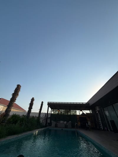 A photograph taken in the city of Ləhiş bağları, Azerbaijan, shows an outdoor swimming pool area with a modern building and some palm trees against a clear blue sky. The pool is filled with turquoise water and has a tiled edge. To the left of the pool, there are three dead palm trees, one of which is significantly taller than the others. Beyond the palm trees, a building with a red tiled roof is partially visible. To the right of the pool, a dark, contemporary building with large windows and a sloped roof is present. An outdoor seating area with wicker furniture is situated under a covered patio structure. A person, appearing to be a male wearing shorts, is visible standing on the deck of the modern building, near a ladder and some cleaning supplies. The overall impression is of a resort or private residence in an arid climate.
