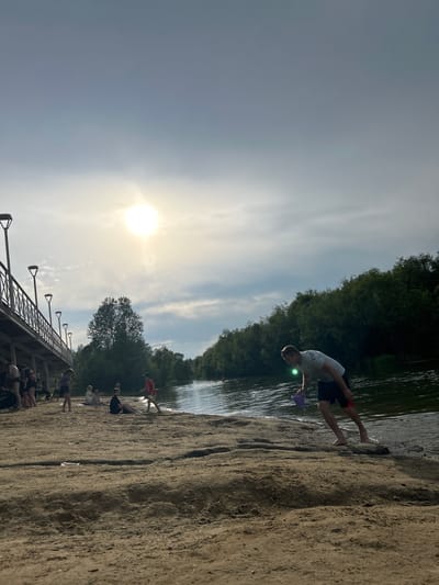 The image depicts a sunny day at a sandy beach next to a body of water, likely a river, as suggested by the presence of trees lining the opposite bank. In the foreground, a young man is bending down at the water's edge, holding a purple bucket and a red object, possibly a shovel. His legs are submerged in the water, indicating he is either entering or exiting the water. In the midground, several people are scattered across the beach. Some are sitting, while others appear to be standing. On the left side of the image, a pedestrian bridge with lampposts runs parallel to the shoreline. The sky is partly cloudy with the sun shining brightly, creating a hazy effect. The overall scene suggests a recreational setting, possibly a public beach. The specific location is identified as Angarsk, Russia, and the scene may be the beach area near the Angara River, a common recreational spot in the city.