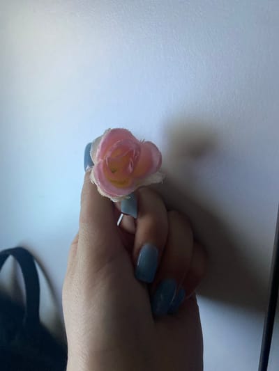 A close-up, high-angle shot captures a human hand gently holding a small, artificial pink flower against a light-colored, textured background. The hand, likely female, features fingernails painted with a glossy, light blue or periwinkle polish. The flower, resembling a miniature rosebud, has soft pink outer petals that transition to a lighter, possibly yellowish, shade at its core. Its petals appear to be made of fabric, showing a slightly frayed edge on some.

The background is an indistinct, granular, off-white or very light grey surface, possibly a wall or tabletop. The lighting is uneven, casting a prominent, dark shadow from the hand and arm across the right side and upper portion of the frame, suggesting an indoor setting with a directed light source. No specific activity is depicted other than the hand holding the flower. There are no discernible location cues within the image itself, nor any visible text, weather conditions, or explicit emotions. The scene takes place in Verkhnevesyoloye, Russia.