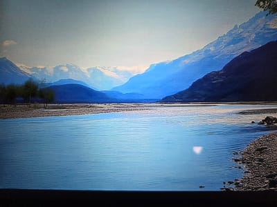 This image, situated in Blainville-sur-Orne, France, depicts a vast natural landscape dominated by a wide body of water and distant mountains. The foreground features a calm, light blue expanse of water, likely a river or lake, with subtle horizontal ripples. A pebbled or rocky shoreline borders the water on both the left and right sides of the frame, with more distinct rocks visible on the right. A distinct, irregular white light patch is present on the water's surface in the lower right quadrant.
In the mid-ground, beyond the immediate water body, a wide, flat expanse of what appears to be a dry, pebbled riverbed or plain stretches across the frame. On the far left of this plain, a cluster of approximately three to five deciduous trees with dark, indistinct foliage is visible.
The background is composed of successive layers of mountains. Closer mountain slopes appear in darker blue-grey tones, particularly prominent on the right side. Further back, multiple ranges recede into progressively lighter blue hues due to atmospheric perspective. The most distant mountains are distinctly snow-capped, exhibiting white peaks under a bright, clear, light blue sky that transitions to a lighter shade near the horizon. No human figures, man-made structures, or specific actions are visible within the scene. The overall environment presents a large, mountainous riverine or lacustrine setting under daylight conditions.