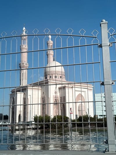 The image shows a view of the Al Noor Mosque in Sharjah, United Arab Emirates, as seen through a metal fence. The mosque features two minarets and a central dome. The building is white with decorative elements. Reflections of water are visible, possibly from the adjacent Khalid Lagoon. The sky is blue.