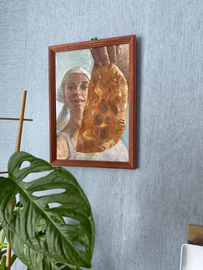 A framed oil painting of a woman wearing a white headscarf and a white top, holding a large, golden-brown, crescent-shaped pastry, is hanging on a light blue textured wall. The painting is framed in a thin, reddish-brown wooden frame. To the left of the painting, a portion of a large, green, fenestrated leaf of a houseplant is in the foreground. A wooden ladder or stand is partially visible behind the plant. The location is the city of Sakmara, Russia.