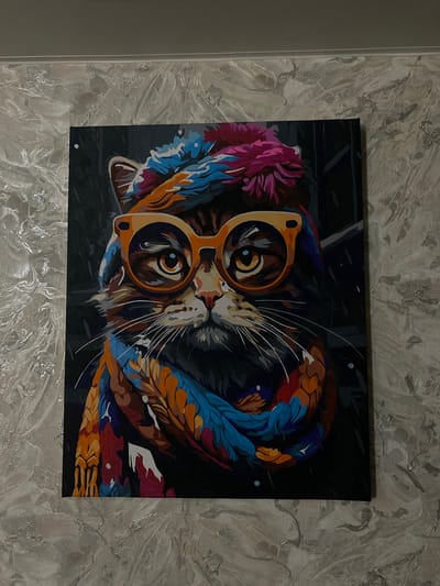 A framed painting of a cat wearing large, orange-rimmed glasses and a colorful scarf and hat is hung on a textured, light-colored wall. The painting depicts the cat's face in a close-up view, with vibrant colors used for its accessories and fur. The background of the painting is dark and indistinct. The wall has a marbled or textured pattern with streaks of white and gray. The image is taken from a slightly angled perspective, with the top edge of the painting slightly higher than the bottom. The lighting appears to be artificial, casting a subtle shadow on the wall to the right of the painting. There is no visible evidence of the city of Sukhoy Ruchey, Russia, in the image.