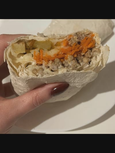 A close-up, eye-level shot captures a hand holding a half-eaten wrap, likely a doner kebab or shawarma, over a white plate. The wrap is made of thin flatbread, and the visible filling includes ground meat, shredded carrots, and sliced potatoes, all bound together with a creamy sauce. The hand is that of a person with dark, glossy nail polish on their fingernails, and the skin appears moisturized. The background is blurred, suggesting an indoor setting. There is no information in the image to suggest a location in Saint-Août, France.