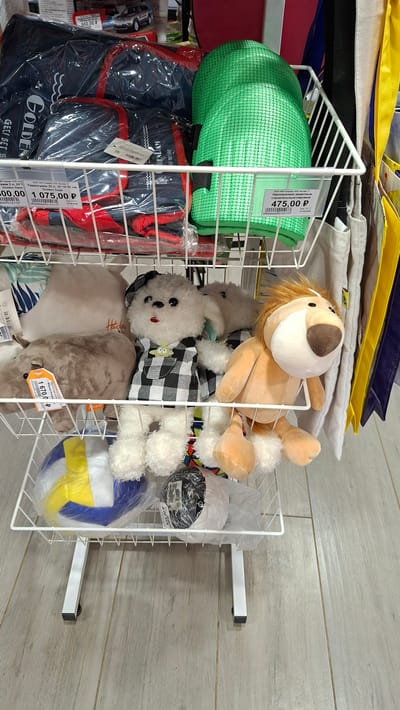 The image displays the interior of a retail store in Nytva, Russia, showcasing various items arranged on white wire shelving units. The foreground features a white wire rack holding several plush toys, including a white dog wearing a checkered outfit, a light brown lion, and a gray hippopotamus. A colorful volleyball is also visible in the lower section of the rack. Behind the plush toys, on a higher shelf, are green knee pads and a blue and red sports bag. The background reveals more merchandise and store fixtures, including yellow and blue shopping bags hanging on the right. The flooring appears to be light-colored wood laminate. The scene is static and depicts a typical retail display.