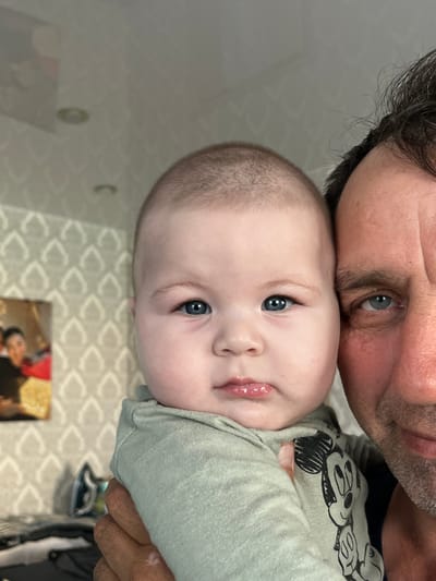 A man and a baby are taking a selfie. The baby is in the foreground, held by the man, and facing the camera. The man is partially visible on the right side of the frame. The baby has blue eyes and is wearing a light green onesie with a Mickey Mouse print. The man has brown hair and stubble and is wearing a dark blue shirt. The background shows a wall with patterned wallpaper and a framed picture. The location is identified as Starobaltachevo, Russia.