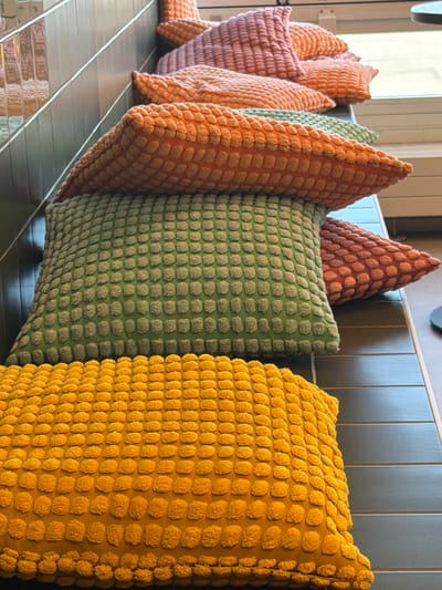 The image displays a stack of textured throw pillows in various colors, including yellow, green, orange, and pink, arranged on a dark, tiled bench. The pillows have a nubby texture, resembling small raised bumps. The background features a tiled wall with a reflective surface, and a window is visible on the right side of the frame. The scene appears to be indoors, possibly in a waiting area or lounge. There are no people or animals present in the image, and no specific location in Malmö-Sturup, Sweden, can be identified.