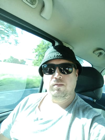 The image shows a man wearing a black bucket hat and sunglasses, sitting in the passenger seat of a car. He is looking at the camera, and his face is illuminated by sunlight. The man is wearing a grey t-shirt. The interior of the car is visible, including the headrest of the seat behind him and a portion of the car's ceiling. Outside the car window, trees and a road can be seen, suggesting the car is in motion. There is no specific indication of the location within the city of Tehov, Czechia.