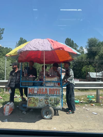 In Ujroo, India, on a sunny day, a mobile food cart is parked on the side of a road with trees and power lines in the background. The cart, labeled "MASALA ROTI," features a colorful, multi-panel umbrella providing shade. Several individuals are present, with three men actively engaged around the cart. One man, wearing a black and white striped shirt and a black cap, holds a water bottle and stands to the left of the cart. Another man, wearing a dark shirt, stands behind the cart, also holding a water bottle. A younger person in a plaid shirt is positioned to the right, appearing to be interacting with the cart. The ground around the cart is littered with various pieces of trash and debris, including plastic bags and paper wrappers. A white and orange shopping bag is visible on the ground to the left of the cart. The overall scene suggests a commercial transaction or preparation of food for sale in an outdoor setting.