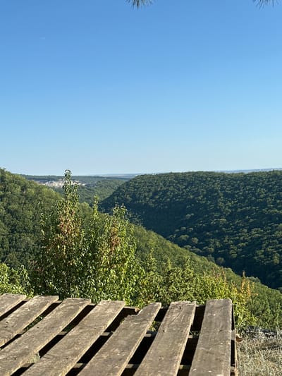 The image shows a scenic view of rolling, tree-covered hills under a clear blue sky. In the foreground, a weathered wooden structure, possibly a bench or a platform made of planks, dominates the lower portion of the frame. The texture of the aged wood, with its visible grain and gaps, suggests it has been exposed to the elements. Beyond this wooden foreground, a lush green valley unfolds, with dense foliage covering the slopes of the hills. In the distance, a lighter patch of land, possibly a quarry or a more open area, is visible, breaking the uniformity of the green landscape. The horizon is marked by a faint line where the hills meet the sky. The scene appears to be a natural landscape, possibly a park or a lookout point, offering a panoramic vista of the surrounding countryside. The location is identified as being in the city of Malozalissia, Ukraine.