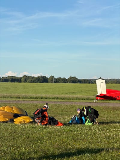 In the city of Limbaži, Latvia, on a grassy field with a clear blue sky and a line of trees in the distance, two individuals are engaged in skydiving preparations. A red and white airplane is parked on a tarmac in the background. In the foreground, a large yellow parachute is laid out on the grass. One person, wearing an orange jumpsuit and helmet with a GoPro camera attached, is kneeling next to a black parachute pack. Another individual, wearing a blue top and purple bandana, is also kneeling, facing away from the camera, with a green and black parachute pack beside them. The scene suggests an active skydiving operation in a rural setting.