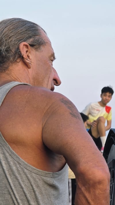 A low-angle shot shows an older Caucasian man with gray hair and a gray tank top, his head tilted back. He has a tattoo on his upper arm. In the background, a young Asian man in a yellow and white shirt appears to be lifting his leg. The location is Unknown, Vietnam.