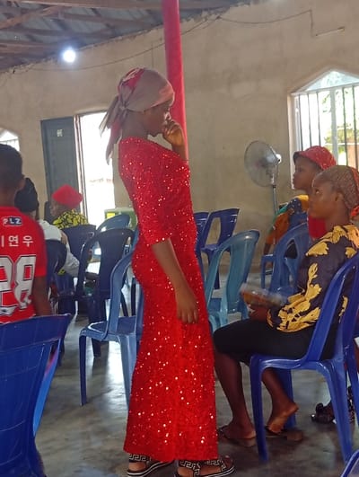 The scene depicts an indoor gathering in Isinyi-Nando, Nigeria. A young adult female stands centrally, facing left, attired in a full-length red sequined dress, a patterned headscarf, and black-and-white patterned sandals. Her right hand is positioned near her mouth. To her right, a second young adult female is seated on a blue plastic chair, wearing a black top with yellow patterns and holding a translucent plastic container. A third female, wearing a red head covering, is partially visible seated further right, adjacent to a grey pedestal fan. Multiple other individuals are partially visible, seated on similar blue plastic chairs throughout the utilitarian room. The environment features light grey, unfinished concrete walls, exposed wooden roof rafters, and a red-painted vertical support pillar. Natural light illuminates the space through windows with metal bars and a doorway. A bare light bulb hangs from the ceiling. A partially visible individual wearing a red and white shirt is seated in the left foreground.