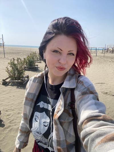 A medium-shot selfie captures a woman on a sandy beach in Ulcinj, Montenegro, under a clear, bright sky. The woman, positioned centrally, has dark hair with distinct magenta highlights on her right side (viewer's left). She possesses light-colored eyes, prominent winged eyeliner, and full lips. She wears a light-toned plaid button-up shirt over a black t-shirt featuring a stylized graphic design of a person. White wired earbuds are visible, connected to a device not pictured. A dark brown strap, possibly from a bag, crosses her right shoulder. She looks directly into the camera with a neutral expression, her head slightly tilted.

The background consists of a wide expanse of sandy beach meeting the sea under a clear blue sky. To the left, a beach volleyball net structure is visible. A row of small, green bushy plants, each contained within a low wooden frame, extends along the sand. Further to the right in the background, several skeletal wooden frames, indicative of beach lounge or sunshade structures, are present. Bright sunlight illuminates the scene, creating a lens flare in the upper left quadrant.