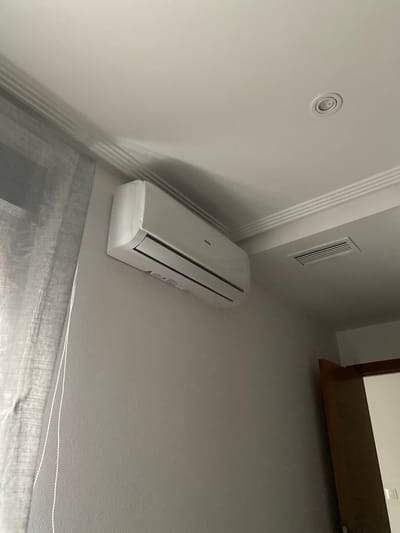 The image shows an interior view of a room with a focus on an air conditioning unit mounted on a white wall. The unit is white and has a sleek, modern design with visible louvers. The wall features decorative moldings, creating a sense of architectural detail. To the left of the air conditioner, there is a recessed circular light fixture on the ceiling. Above and to the right of the AC unit, there is a rectangular ventilation grate. Further to the right, a dark wooden shelf is visible, extending from the wall. The lower part of the image shows a textured gray curtain or blind. The scene appears to be well-lit, suggesting it could be daytime, though no windows or exterior views are clearly visible. The overall impression is of a clean and contemporary living space. The text "Haier" is visible on the air conditioning unit. The location is San Pedro del Pinatar, Spain.