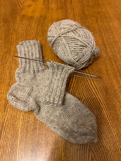 The image displays a pair of partially knitted gray socks, a ball of gray yarn, and knitting needles on a wooden surface. The knitting needles are positioned within the partially completed sock, indicating that the knitting process is in progress. The wooden surface appears to be a table with a visible wood grain pattern. The lighting suggests an indoor setting, possibly a home environment. There are no people present in the image. The scene is static and does not depict any specific action or interaction beyond the implied activity of knitting. The image is taken from a top-down perspective. The specific location of Aginskoye, Russia is not visually discernible from the provided image content.