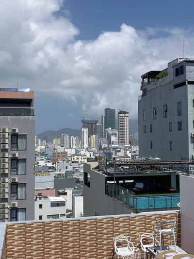 An aerial view captures an urban cityscape in Ngũ Hành Sơn Ward, Vietnam, from a high-rise rooftop perspective under a partly cloudy sky. In the foreground, a tiled rooftop terrace features a partial view of a small white table and three white metal chairs, two with distinctive arched backs. To the immediate left, a multi-story grey building presents a series of vertically stacked external air conditioning condenser units on its facade, with multiple windows visible. To the right, another multi-story grey building showcases a rooftop swimming pool with a glass safety barrier, beside which a dark lounge chair with a white towel is visible. Above the pool, an additional rooftop level is present with dark railings and sparse greenery.
Beyond these immediate structures, a dense urban landscape unfolds with numerous multi-story buildings of varying heights and architectural styles, predominantly white and grey. A yellow "S HOTEL" sign is visible on one of the mid-level buildings. In the distant background, several taller high-rise buildings dominate the skyline, including some under active construction with visible cranes and a distinctive green-colored high-rise. Faint mountain ranges are discernible on the distant horizon. The sky exhibits a mix of bright blue patches and large, white cumulus clouds, with darker cloud formations appearing towards the distant horizon. No individuals or specific actions are visible within the frame.