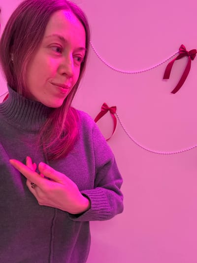 A female subject is captured from the chest up, positioned in a setting illuminated by vibrant pink light. She has light brown hair, appearing reddish-purple due to the lighting, and is dressed in a grey turtleneck sweater. Her gaze is directed to the right of the frame, accompanied by a subtle smile. Her left hand is raised, with the index finger extended and pointing right, while the other fingers are curled. A ring with a dark, possibly red, stone is visible on her left ring finger. The background consists of a solid wall adorned with several small red ribbon bows and strands of white beaded chains, creating a decorative arrangement. This scene is located in the city of Novosergievka, Russia.