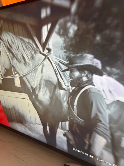 The image depicts a still from a video game, likely Red Dead Redemption 2, given the aesthetic and on-screen text. The scene is monochromatic and features a man in Western attire, including a hat and suspenders, standing next to a horse in what appears to be a stable or barn. The man is looking to the left, and the horse is facing forward. The environment is dimly lit, with shadows cast by wooden structures. On-screen text in Russian, "ФОТОРЕЖИМ" (Photo Mode) and "ВЫБРАТЬ" (Select), is visible at the bottom right of the image, confirming its origin as a video game interface. There is no indication of the city of Geamăna, Moldova, being present in this image.