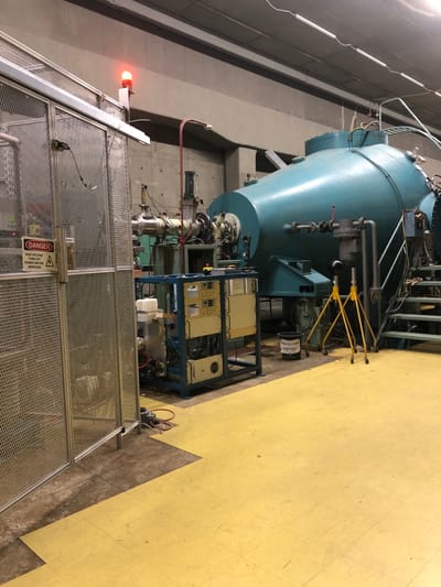 The image shows the interior of a large industrial facility, likely a laboratory or research center. It is located in Agia Paraskevi, Greece, at coordinates 37.998 latitude and 23.82 longitude. The main subject of the image is a large, blue, cylindrical object, possibly a vacuum chamber or a reactor. This chamber is positioned in the center of the room and connected to various piping and instrumentation. To the left of the chamber, there is a metallic cage with a warning sign that reads "Danger - High Voltage, Turn off Power Before Servicing". The cage surrounds a complex piece of equipment, possibly a control panel or a power supply unit. The floor is a bright yellow, and there are several yellow warning signs scattered around the room. The overall impression is of a well-equipped and potentially hazardous research environment.