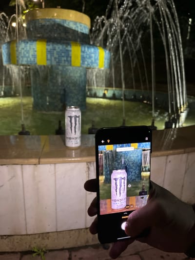 The image captures a nighttime scene in Veliki Preslav, Bulgaria, focusing on a person photographing a beverage can. In the foreground, a right hand holds a dark-colored smartphone, with its screen displaying the camera interface. The phone's camera is actively focused on a white Monster Energy Ultra Zero Sugar can, which stands upright on a beige tiled surface forming the edge of a water fountain. The phone screen shows "FLASH ALWAYS ON" and a "2.5x" zoom indicator.
The water fountain, located in the mid-ground, features multiple tiers adorned with blue mosaic tiles and yellow vertical stripe accents. Water actively cascades and sprays from several jets within the fountain, pooling in a basin where the water appears greenish due to ambient lighting. Several dark, small nozzles are visible within the water. The background consists of dark areas, with indistinct light sources. The ground beneath the smartphone consists of light-colored paving or tiling, with a small patch of green vegetation visible on the far left.