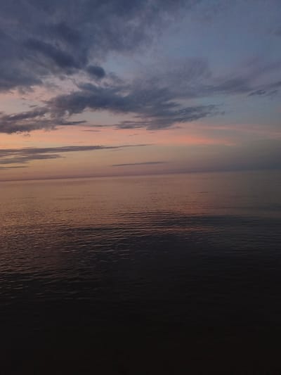 This is an image of the Baltic Sea at dusk, as seen from the coast in Apšuciems, Latvia. The sky is filled with scattered clouds, tinged with hues of pink and purple from the setting sun. The sea is calm, reflecting the colors of the sky. The lower portion of the image is dark, suggesting the absence of artificial light and the deepening twilight. There are no people or structures visible in the scene, creating a serene and natural landscape.