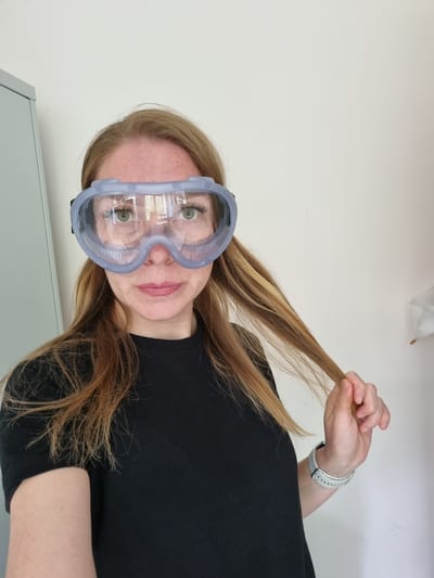 A woman with auburn hair and green eyes, wearing clear safety goggles and a black t-shirt, poses for a selfie in a room with a light-colored wall. She is holding a strand of her hair in her right hand, which is also adorned with a white watch. To her left, a portion of a gray cabinet is visible. The room appears to be indoors, with no clear indicators of its specific location or any connection to the city of Kemul, Russia.