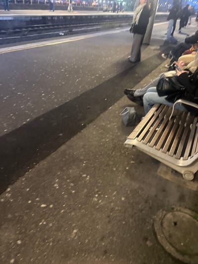 The image depicts an outdoor scene, likely at a train or tram station platform in Molsheim, France, during the evening or night. A person is seated on a wooden slatted bench with metal supports on the right side of the frame. They are wearing light blue jeans and black shoes, with a dark bag resting on the ground near their feet. Their upper body is partially visible, and they appear to be engaged with a phone. To the far right, a circular metal manhole or drain cover is visible on the pavement.

Further left, another individual stands, partially visible from the waist down, dressed in a dark coat and trousers. In the blurred background on the far left, there are indistinct figures and bright, artificial lights, suggesting a public area adjacent to train or tram tracks and a platform edge. A long, distinct dark shadow stretches diagonally across the dark asphalt or concrete ground from the upper right. The ground itself is textured with scattered light specks. The scene conveys an atmosphere of urban transit and waiting. No text is visible.