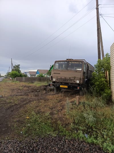 This image depicts a dusty, rural roadside scene in Shakhtinsk, Kazakhstan. The main subject is a weathered, brown KAMAZ truck parked precariously close to a wooden building and an overgrown bush. The truck's windshield has two stickers with the KAMAZ logo. A yellow license plate is visible on the front bumper, displaying the characters "KZ F 8594 09".
The setting appears to be a residential area, with a wooden fence running along the properties. Several houses with colorful roofs are visible in the background, along with trees and a satellite dish. A power line pole with multiple wires runs vertically along the right side of the frame, stretching into the cloudy sky. The ground is covered in dry grass, dirt, and small rocks. Some litter is scattered on the ground.
The sky is overcast with heavy gray clouds, suggesting a possibility of rain or a general dull day. The time of day is likely late afternoon or early evening, given the subdued lighting. There are no people visible in the frame. The overall mood is one of stillness and perhaps neglect, with the truck appearing to be out of commission or temporarily parked. The environment suggests a relatively remote or less developed area.